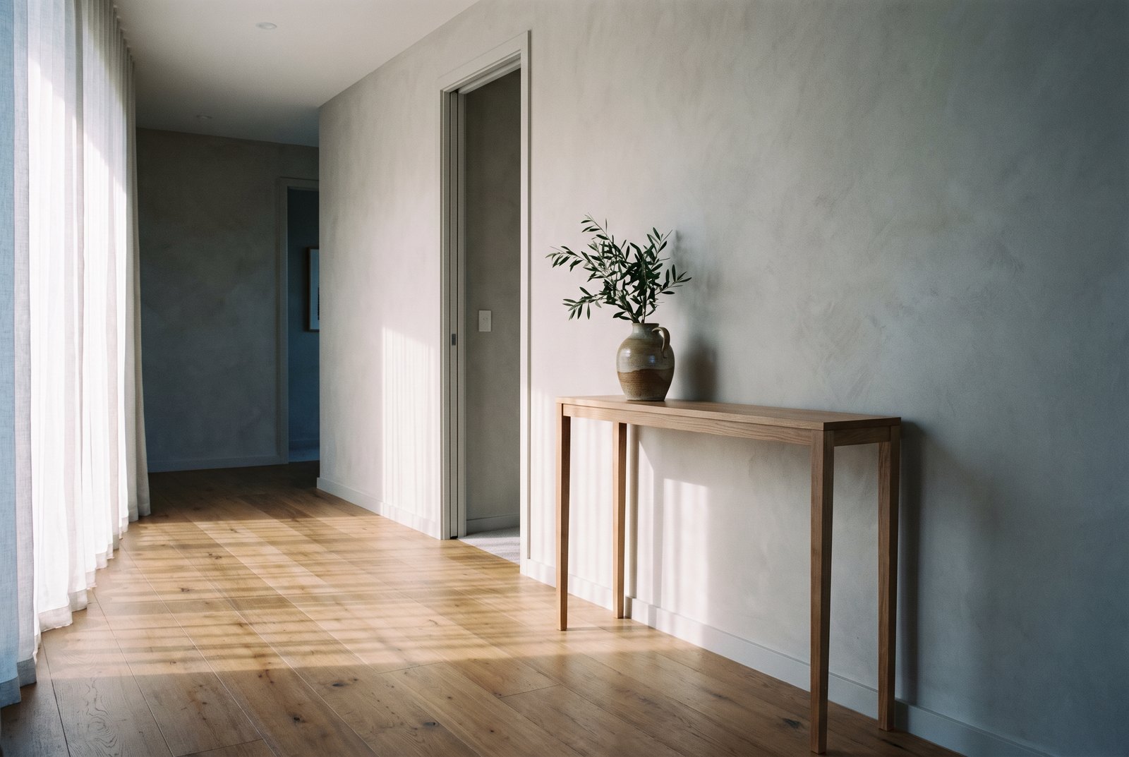 A quiet hallway in soft morning light, warm timber floor, plaster wall, a console table with an olive branch